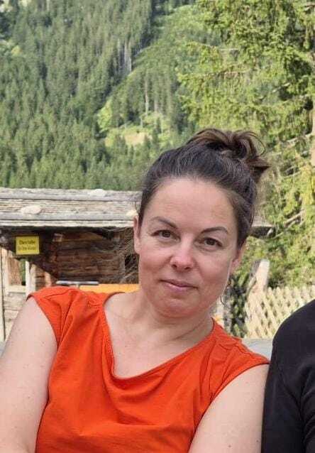 Frau mit Dutt trägt ein oranges Oberteil, sitzt vor alpenländischer Landschaft mit Bäumen und Hütten im Hintergrund.