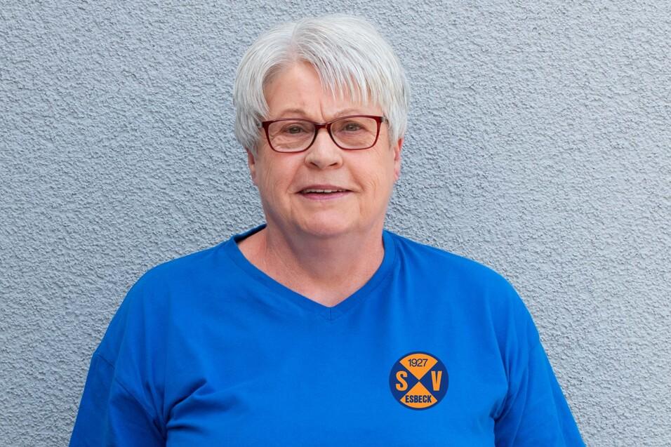 Porträt einer lächelnden Frau mit grau-weißen Haaren in einem blauen T-Shirt vor einer grauen Wand.
