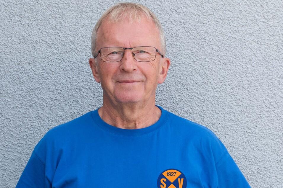Älterer Mann mit Brille trägt ein blaues T-Shirt mit dem Logo eines Sportvereins vor einer grauen Wand.