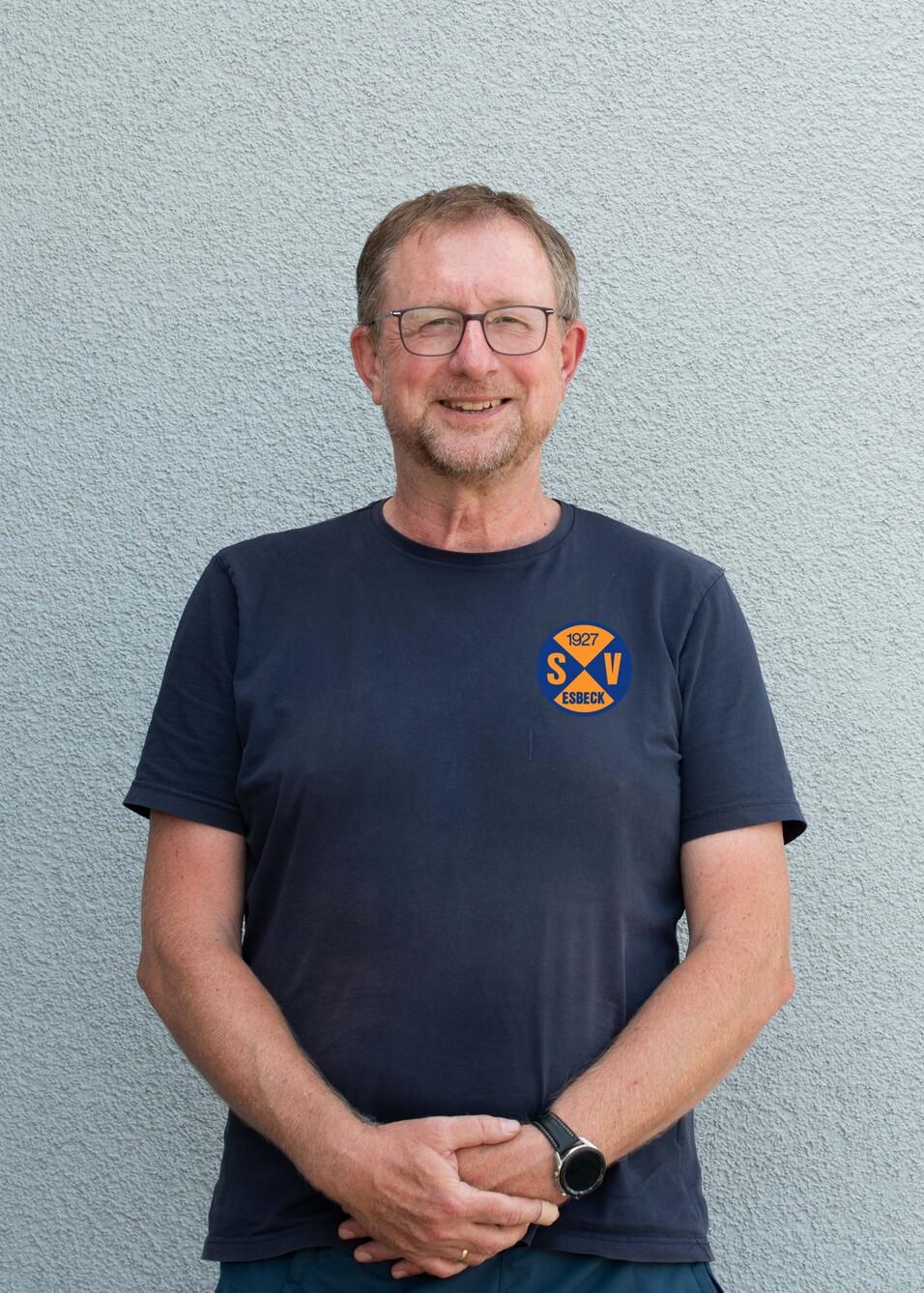 Porträt eines lächelnden Mannes mit Brille, der in einem T-Shirt mit Vereinslogo vor einer grauen Wand steht.