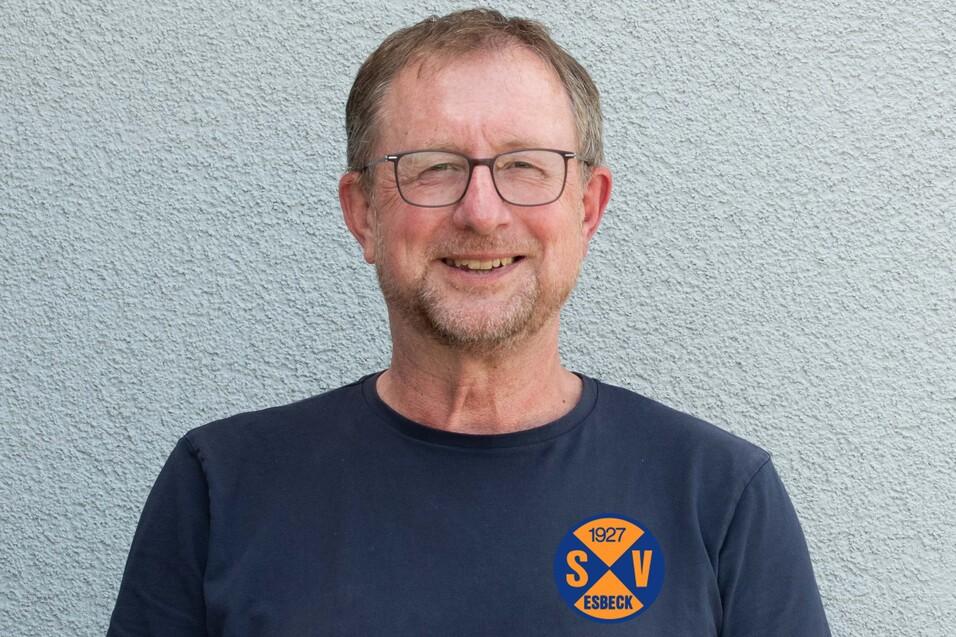 Porträt eines lächelnden Mannes mit Brille, der in einem T-Shirt mit Vereinslogo vor einer grauen Wand steht.