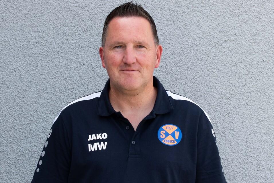 Mann in dunkelblauer Sportkleidung mit Vereinslogo steht vor einer grauen Wand.