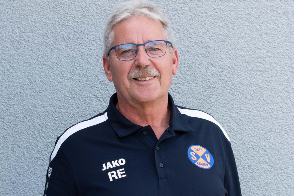 Männlicher Trainer in einem schwarzen Poloshirt mit Vereinslogo steht vor einer grauen Wand.