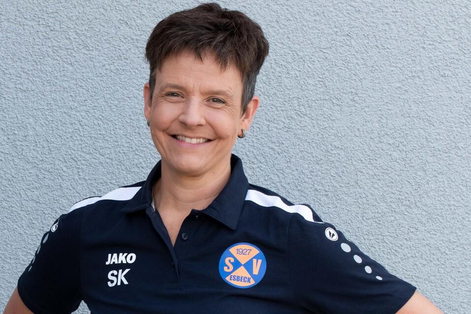 Frau mit kurzem Haar trägt ein dunkles Sportshirt mit Vereinslogo und steht lächelnd vor einer grauen Wand.