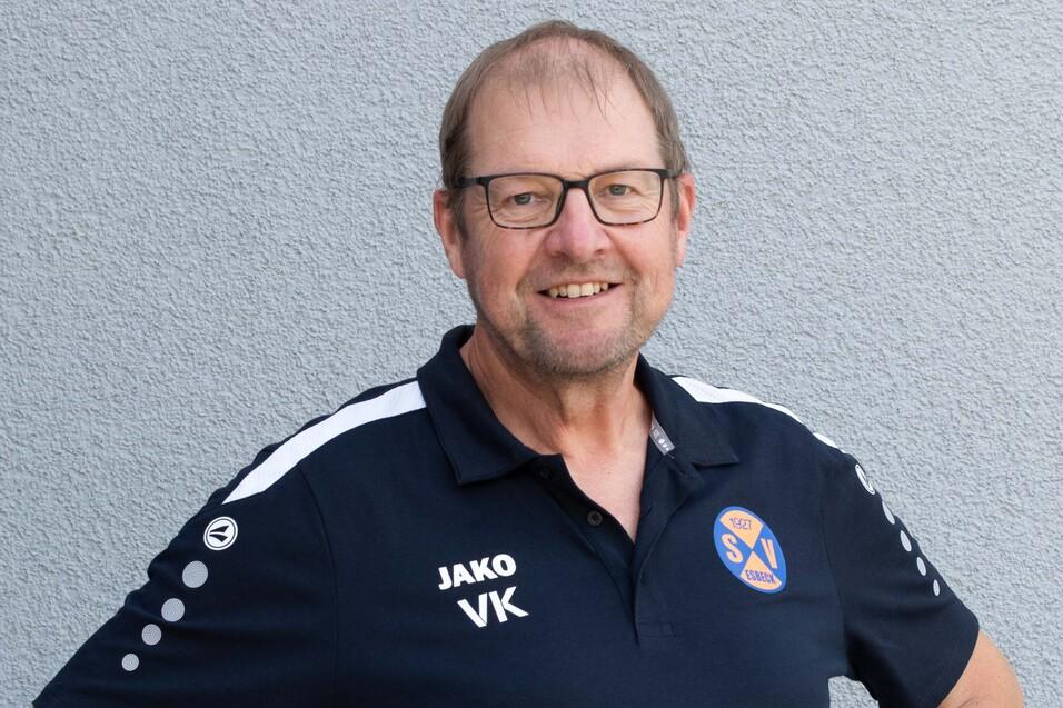 Portrait eines lächelnden Mannes in einem schwarzen Sporttrikot mit Brille vor grauem Hintergrund.