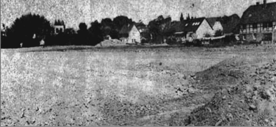 Schwarz-Weiß-Foto einer leeren, unebenen Fläche mit Gebäuden im Hintergrund und Bäumen am Rand.