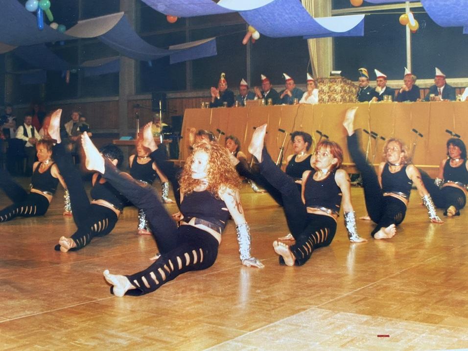 Tanzgruppe in Schwarz führt synchronisierte Bewegungen auf einer Bühne mit Zuschauern im Hintergrund aus.