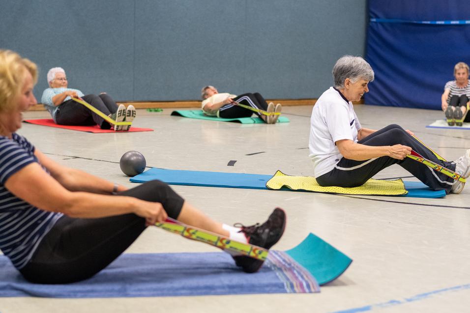 Senioren führen Rückenübungen mit Widerstandsbändern auf Matten in einer Sporthalle durch. Diese sind in verschiedenen Farben.