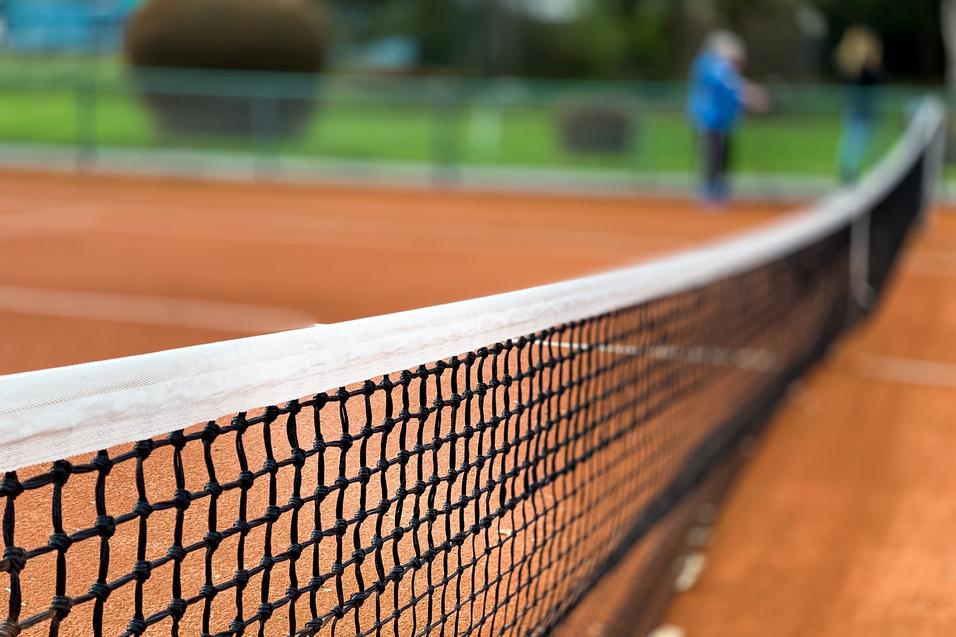 Nahaufnahme eines Tennisnetzes auf einem roten Sandplatz, Hintergrund unscharf mit grünen Bäumen.